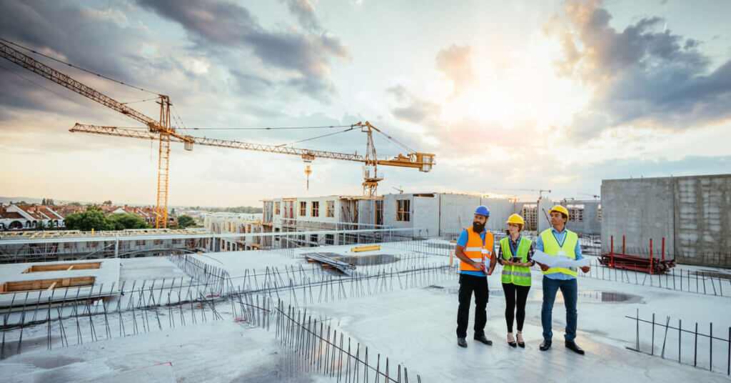 Three colleagues in safety gear reviewing plans on the top of a building under construction.