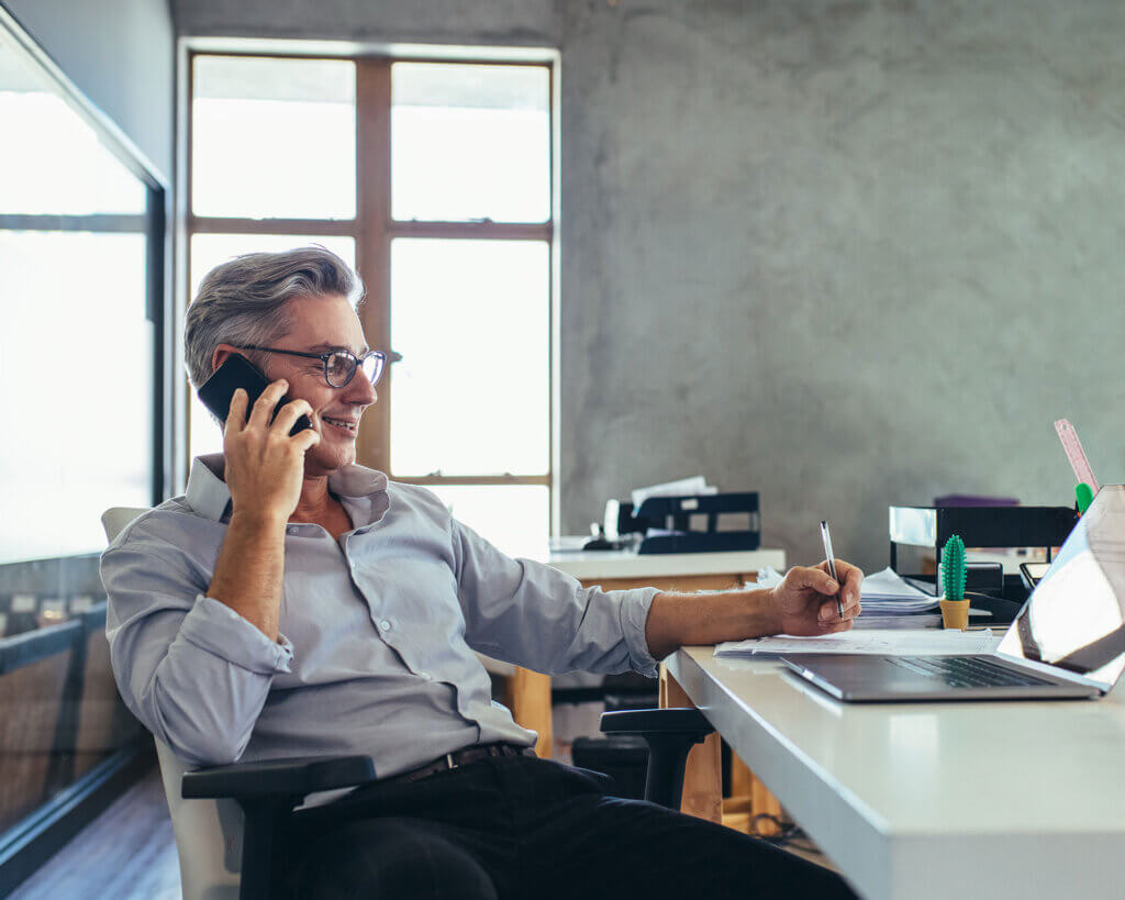 Businessman sitting in an office talking on a cell phone with laptop in front of him.