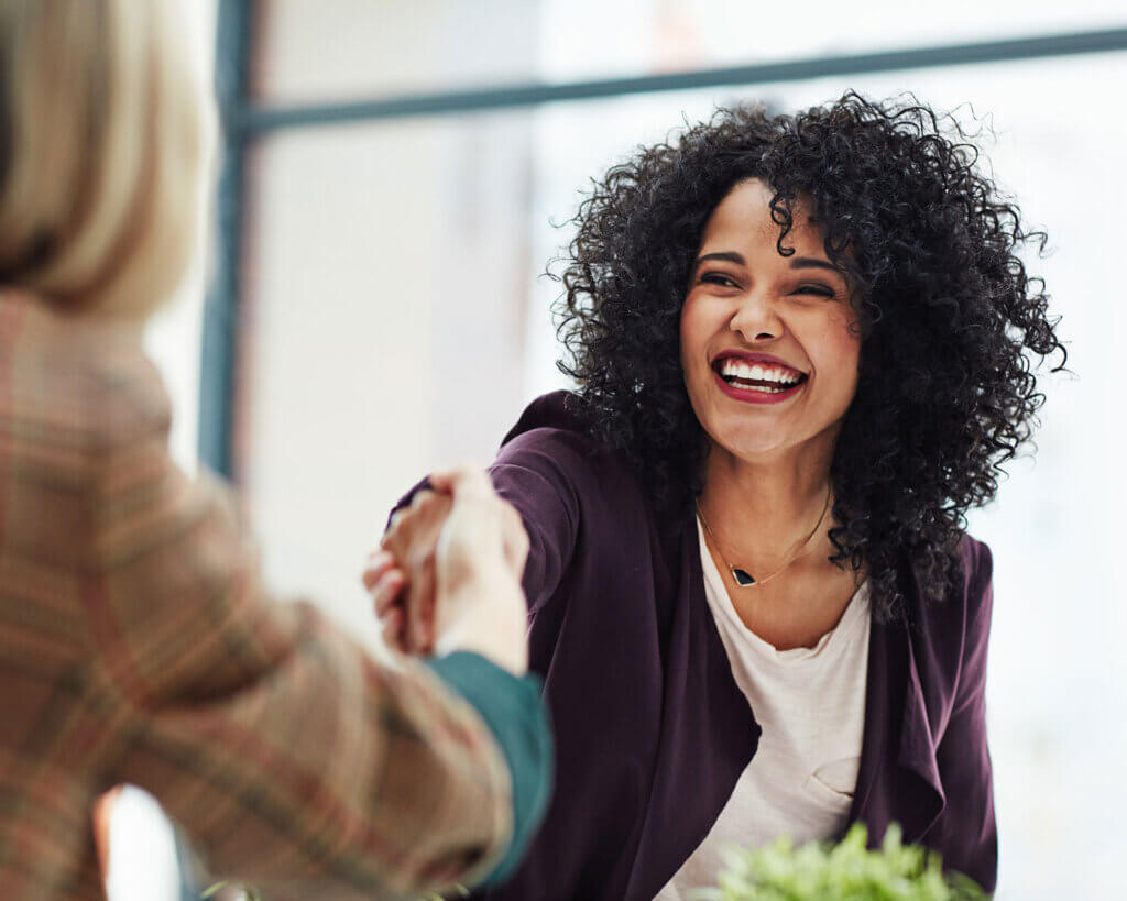 Smiling businesswoman wearing a purple sweater and white shirt shaking hands with a client.