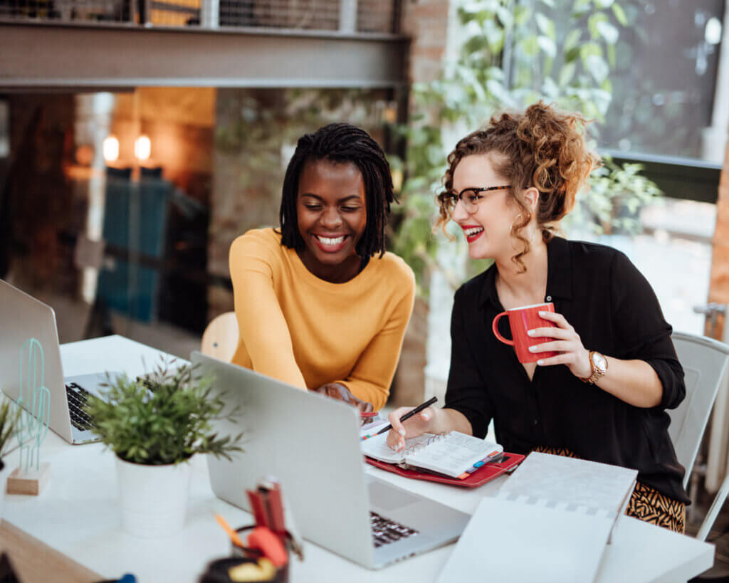 two women laughing together while looking at laptops