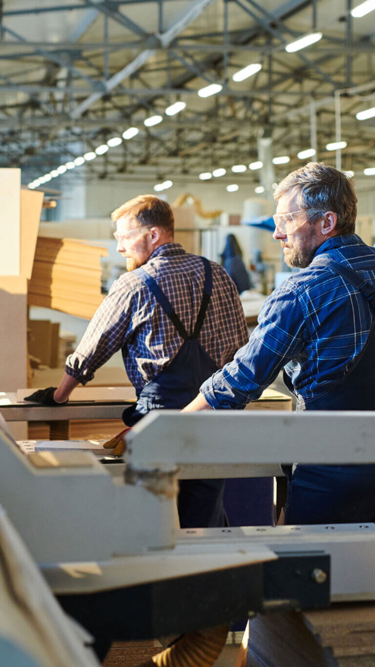 Two woodshop workers at the table saw cutting large pieces of wood.