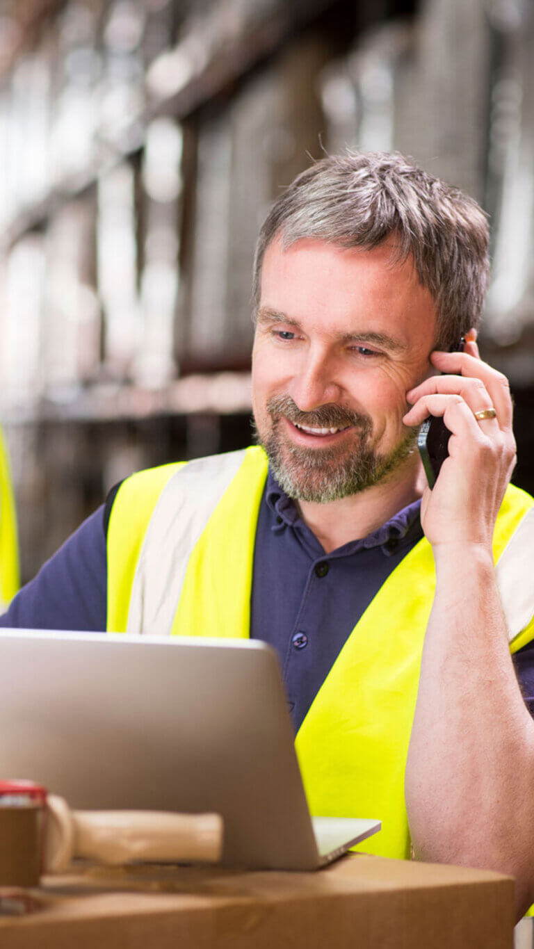 Warehouse manager on phone in front of laptop, inside warehouse.