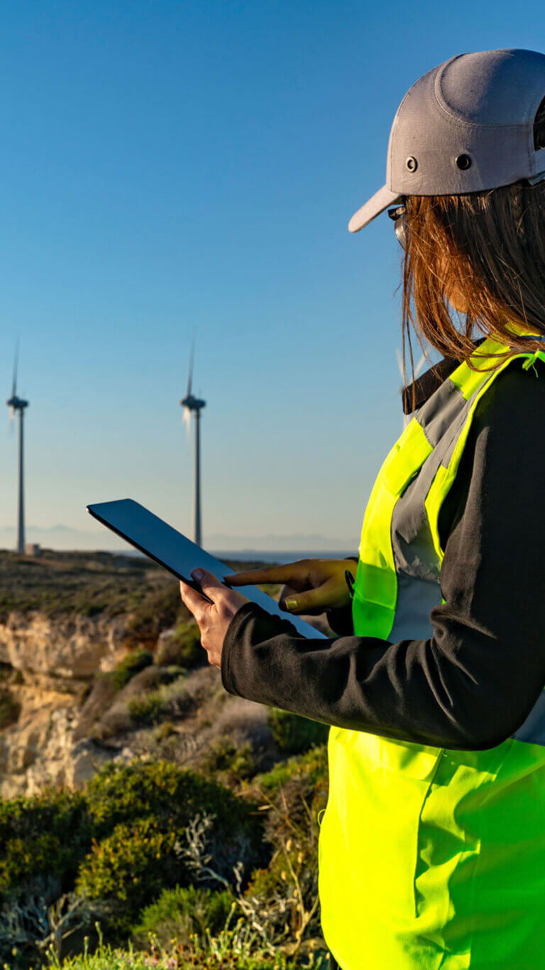 Wind turbine engineer reviewing renewable energy data on her tablet, standing in front of wind turbines.
