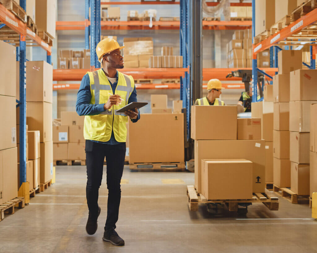 Male worker wearing hard hat holding digital tablet walking through retail warehouse.